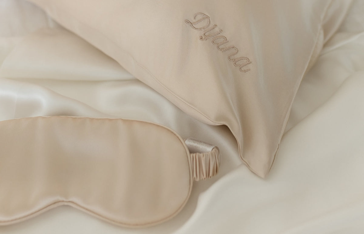 A corer of silk pillowcase in beige with embroidery "Dijana' tone-on-tone in matching color thread and silk sleep mask next to it.