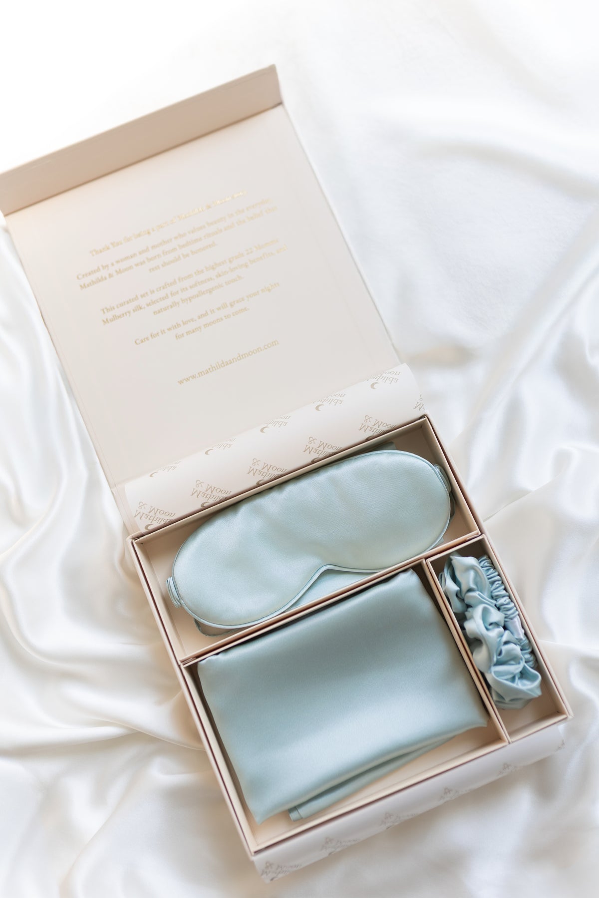 Silk Set of light blue eye mask, folded pillowcase, and hair ties in a gift box on a white fabric background