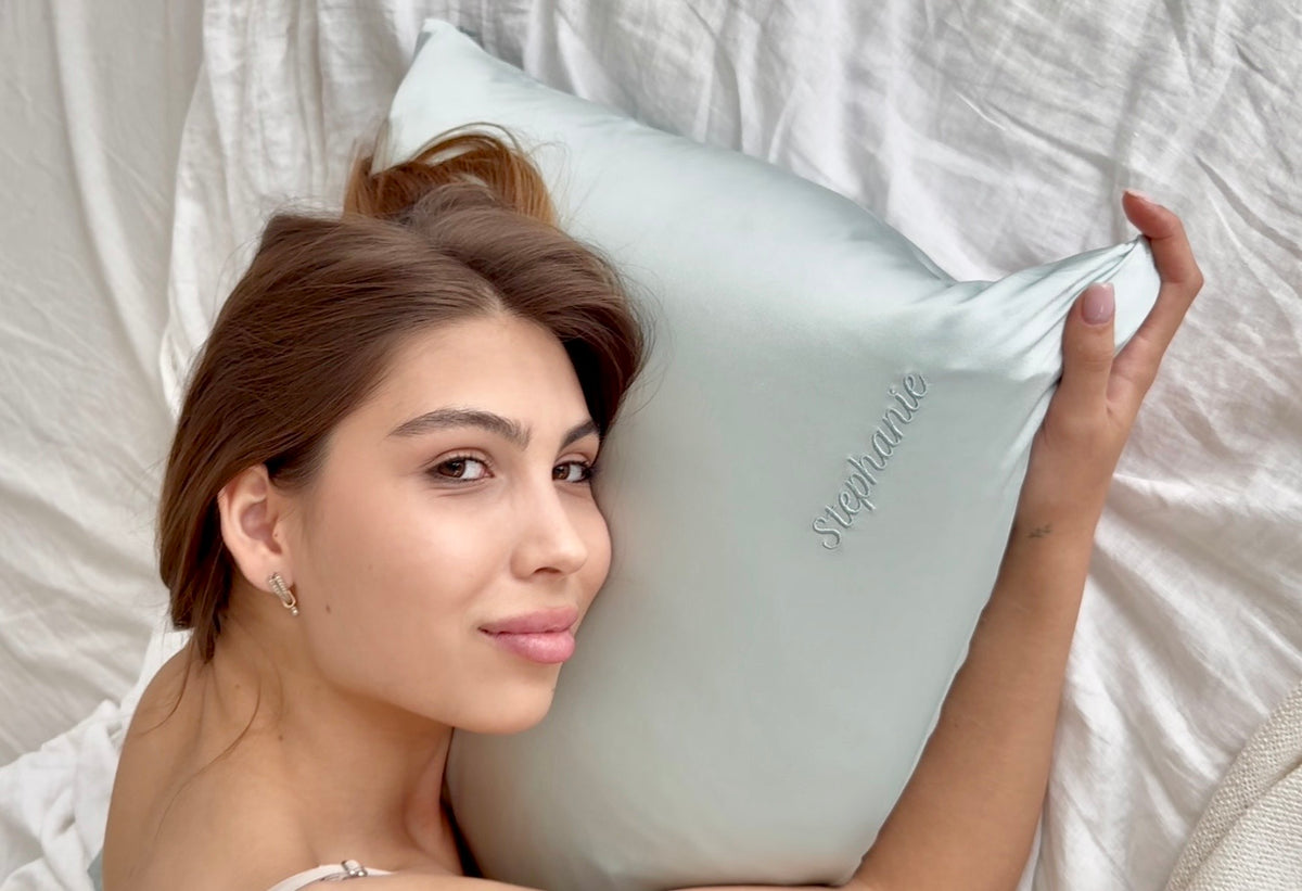 A woman lying in bed with a pastel blue pillow, holding one corner. The pillow has the name 'Stephanie' embroidered on it in a matching pastel blue thread.