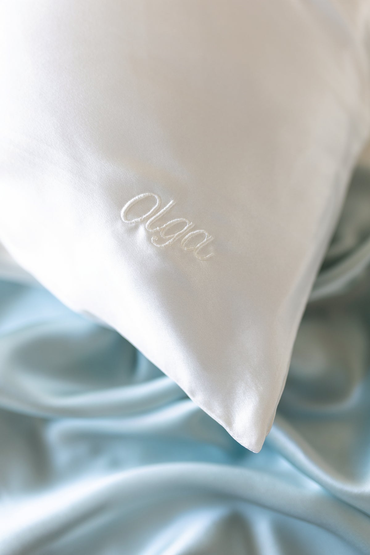 A close-up of a white silk pillowcase with the name 'Olga' embroidered on tone-on-tone white script, resting on a light blue silk sheet.