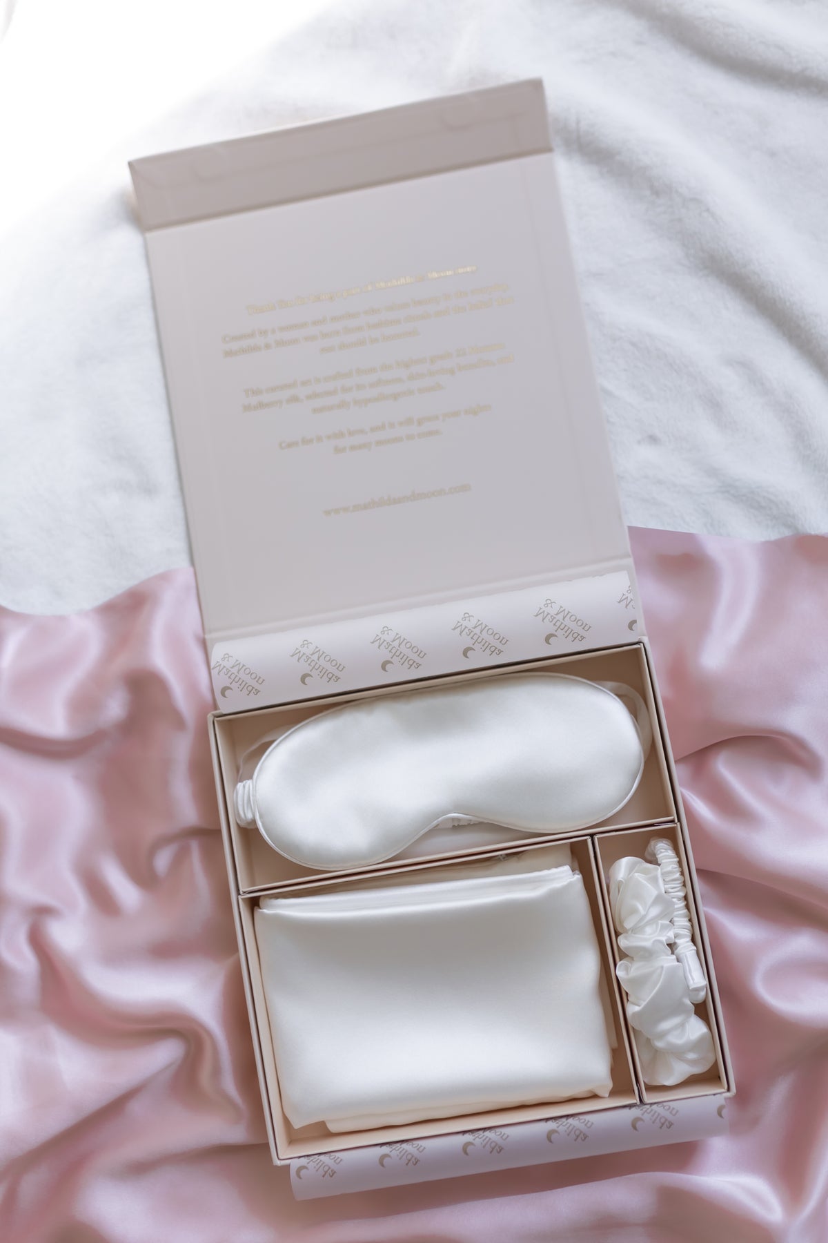 Set of silk white eye mask, pillowcase, and two scrunchies in a decorative box on pink fabric.